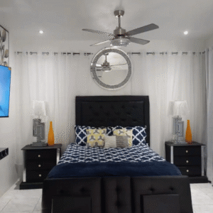 Serenity Vibes apartment bedroom featuring a stylish black headboard, blue patterned bedding, ceiling fan, wall mirror, bedside lamps, and floor-to-ceiling curtains