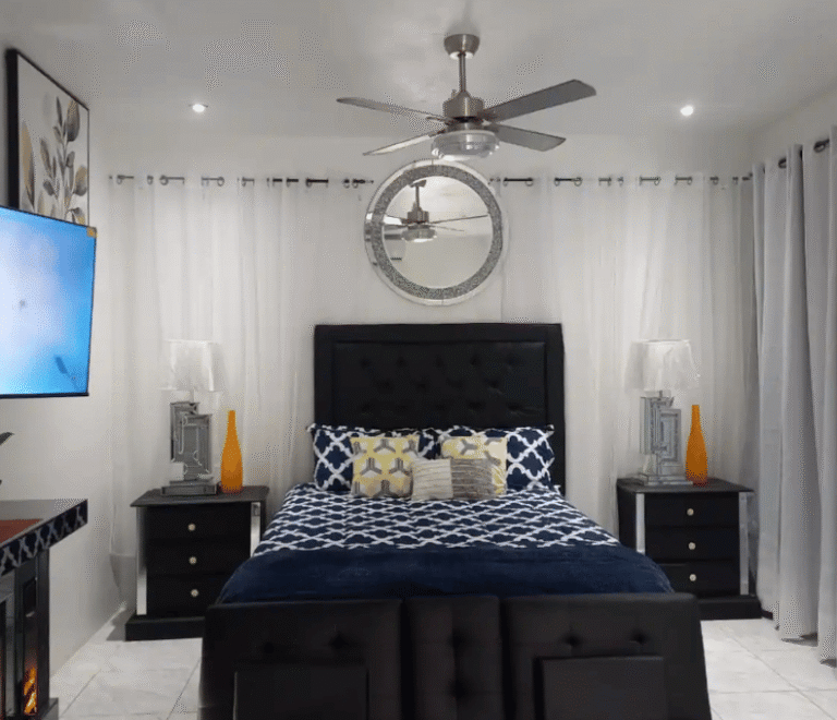 Serenity Vibes apartment bedroom featuring a stylish black headboard, blue patterned bedding, ceiling fan, wall mirror, bedside lamps, and floor-to-ceiling curtains