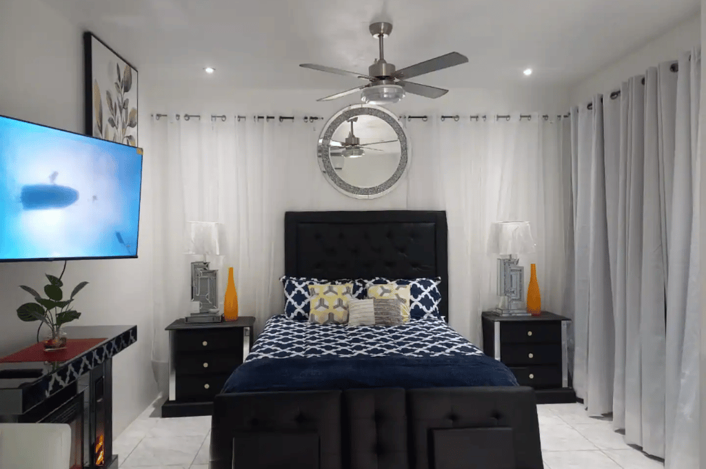 Serenity Vibes apartment bedroom featuring a stylish black headboard, blue patterned bedding, ceiling fan, wall mirror, bedside lamps, and floor-to-ceiling curtains